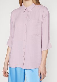 Woman wearing a loose light purple button-up shirt with three-quarter sleeves and a chest pocket, paired with shiny light blue pants.
