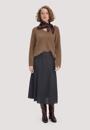 Femme portant un pull en maille marron, une jupe midi noire, une écharpe marron foncé nouée autour du cou et des bottines à talons marron foncé, debout devant un fond uni.
