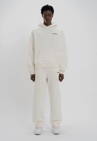 Κρέμα φούτερ και ταιριαστά παντελόνια φόρμας από μαλακό ύφασμα, με μπροστινή τσέπη και λογότυπο. Ο σχεδιασμός είναι χαλαρός.