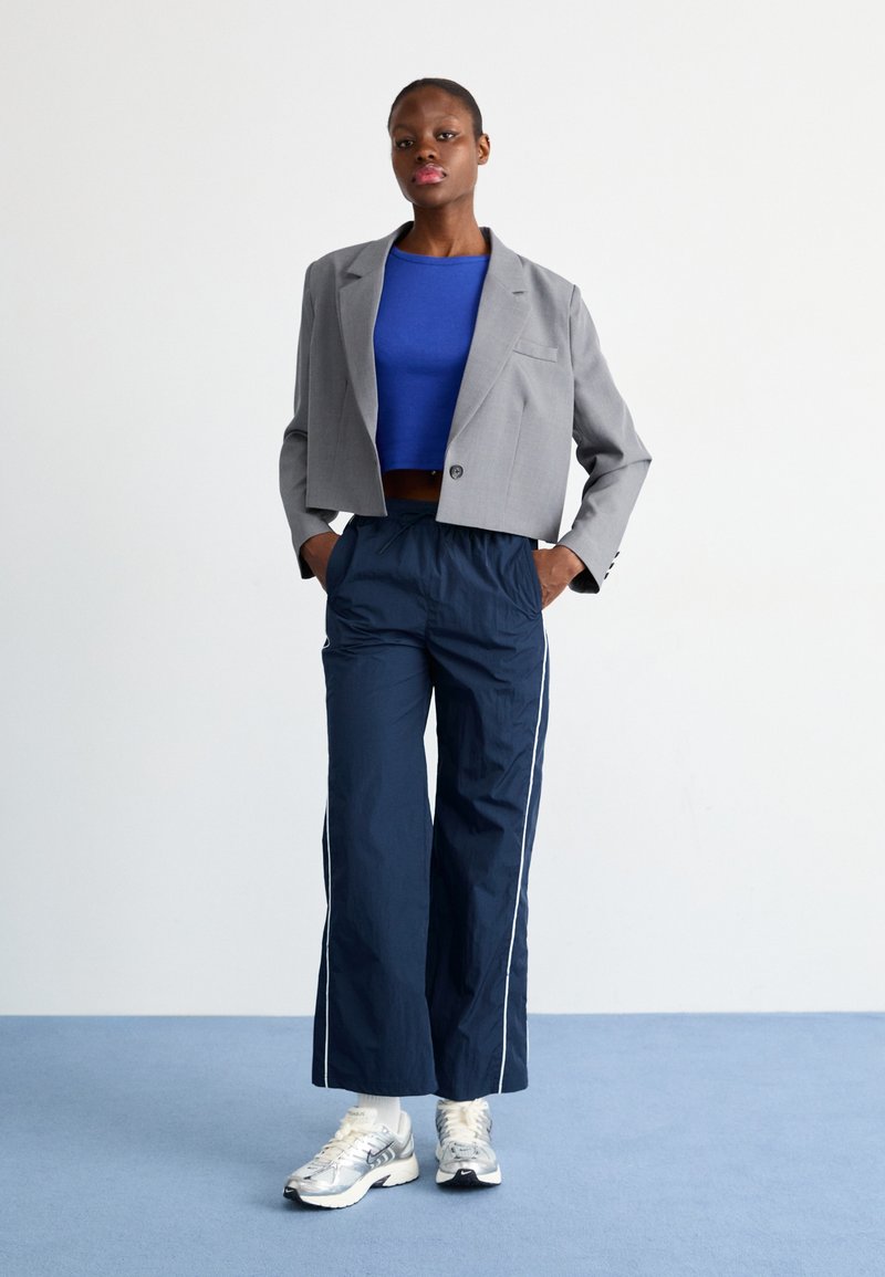 Grijze cropped blazer over een blauwe aansluitende top, gecombineerd met marineblauwe wijde broek met witte zijkanten en zilveren sneakers.