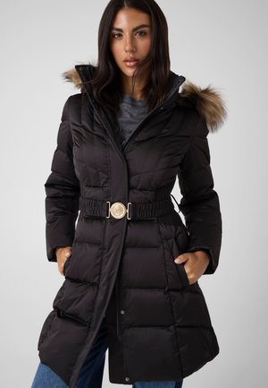 Mujer con un abrigo de invierno negro acolchado, con capucha adornada con piel, cintura ceñida con cinturón y las manos en los bolsillos laterales, sobre un fondo liso.