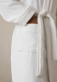 Weißer Bademantel aus weichem, strukturiertem Stoff. Verfügt über einen gebundenen Bund und eine große Fronttasche auf der linken Seite. Einfaches und minimalistisches Design.