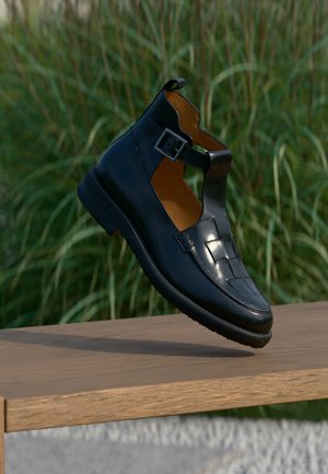 Schwarzer Leder-Loafer mit Schnallenriemen schwebt über einer Holzbank mit grünem Gras im Hintergrund.