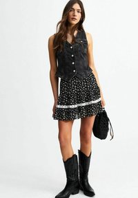 Chaleco de mezclilla negro con un diseño ajustado, combinado con una falda acampanada negra con estampado floral y un borde de encaje blanco. Botas de vaquero negras y bolso.