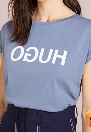 Kobieta ubrana w jasnoniebieską koszulkę z białym, odwróconym napisem "HUGO" oraz ciemne, z wysokim stanem spodnie z widocznym białym przeszyciem.