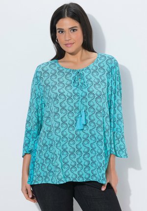 Kobieta ubrana w luźną turkusową bluzkę w czarny wzór pętli, z wiązaniem przy dekolcie ozdobionym frędzlami oraz czarne spodnie na jednolitym białym tle.