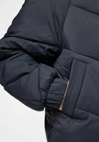 Námořnická pufovaná bunda s tlustým prošívaným designem. Obsahuje boční kapsu a elastické manžety pro těsné usazení a teplo. Hladký, matný povrch.