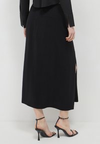 Jupe noire midi avec fente latérale, fabriquée dans un tissu lisse. Associée à des sandales à talons aiguilles noires avec des brides à la cheville.