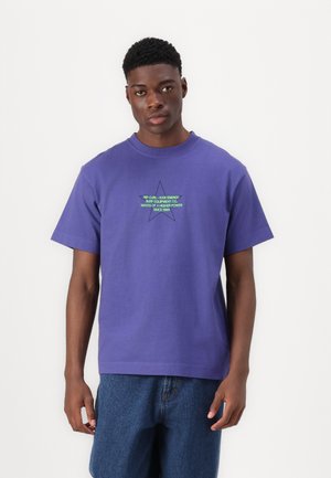 Camiseta de algodón morada de manga corta, con un estampado gráfico de una estrella verde en el pecho. Combinada con pantalones cortos de denim azules.