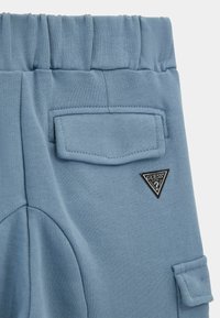 Pantaloni de trening bleu deschis cu un tiv texturat, buzunare laterale și un buzunar aplicat cu un logo triunghiular negru. Textură netedă.