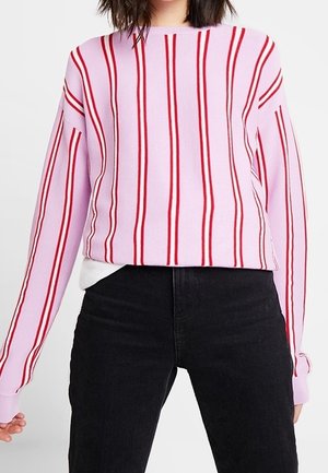 Kvinde iført en pink langærmet trøje med lodrette røde striber, kombineret med sort høj talje jeans.