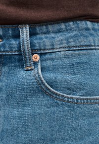 Κοντινή λήψη τσέπης μπλε τζιν και θηλιάς ζώνης με χάλκινη λεπτομέρεια καρφιτσών, συνδυασμένα με καφέ υφασμάτινο πουκάμισο μέσα στην μπλούζα.