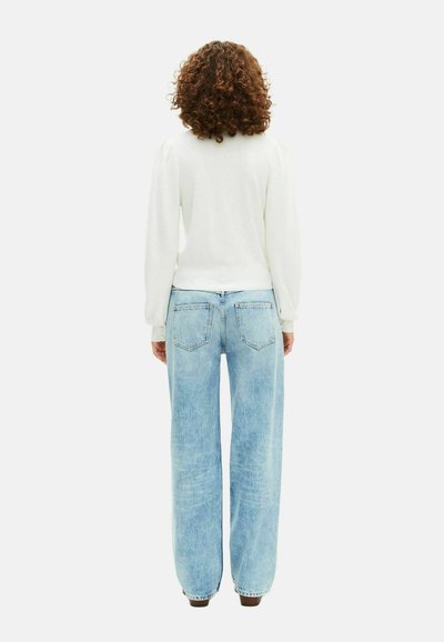 Personne avec des cheveux bouclés portant un haut blanc à manches longues et un jean large bleu clair, faisant face à l'arrière sur un fond uni.