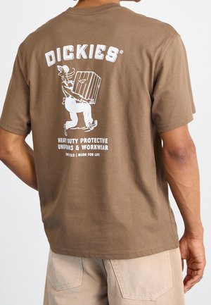 Uomo che indossa una maglietta marrone Dickies con un cartone animato bianco che porta una scatola e testo sulle uniformi protettive resistenti sul retro.