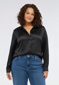 Camicia in satin nero con maniche lunghe, collo e piccole decorazioni a pois argentati. Abbinata a jeans blu a vita alta.