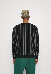 Černý svetr s vertikálními bílými pruhy, s žebrovanými manžetami a lemem. Model má na sobě zelené kalhoty, což ukazuje zadní pohled na oděv.