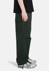 Groene cargo broek met meerdere zijzakken, relaxed fit, en een trekkoord bij de enkels, gecombineerd met zilveren sneakers.