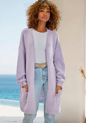 Lila langer Cardigan mit lockerer Passform, gerippten Bündchen und zwei vorderen Taschen, getragen über einem weißen Oberteil und hellblauen Jeans. Weiche Textur.