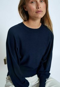 Dunkelblauer Langarmpullover aus weichem Strickmaterial, mit rundem Ausschnitt und geripptem Saum. Getragen mit hellen Hosen.
