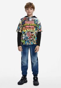 Maglietta a maniche corte con grafica colorata di un supereroe Marvel, con maniche lunghe nere sotto. Abbinata a pantaloni jogging in denim blu e sneaker nere.