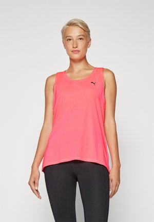 Junge Person mit kurzem blonden Haar, die ein hellpinkfarbenes, ärmelloses Sportoberteil und schwarze Leggings trägt und vor einem schlichten Hintergrund steht.