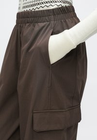 Hnedé ľahké nohavice s elastickým pásom, bočnými vreckami a nákladným vreckom na stehne. Hladká textúra a jednoduchý dizajn.