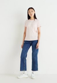 T-shirt rosa chiaro a maniche corte con scollo rotondo, abbinato a jeans blu a gamba larga. Il modello indossa sneakers bianche con dettagli chiari.