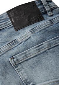I jeans in denim azzurro chiaro presentano un'etichetta in pelle nera testurizzata con testo in rilievo. Sono visibili diversi taschini nella parte posteriore.