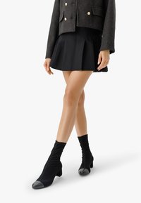 Femme portant une veste courte noire, une mini-jupe plissée noire et des bottines à talons noires avec des embouts arrondis.