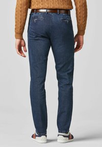 Jeans en denim bleu foncé à coupe slim, avec deux poches arrière et une ceinture en cuir marron. Les revers sont légèrement retroussés, dévoilant des chaussures.