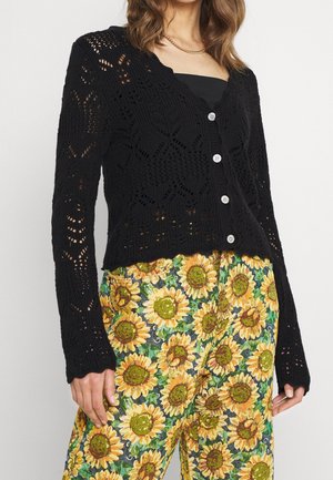 Femme portant un cardigan en tricot noir à boutons blancs sur un haut noir et un pantalon taille haute orné de grands tournesols jaunes.