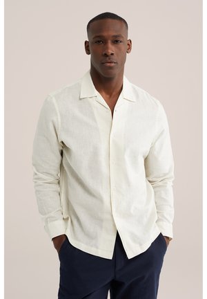 Mees, kes kannab kahvatuvalget pikade varrukatega nööbitavat särki avatava kraega ja tumesiniseid pükse, seisab taskutes kätega ühevärvilise tausta ees.