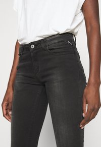 Musta denim farkut, joissa on kapea leikkaus, vakiomallinen viiden taskun malli ja metallinen nappikiinnitys vyötäröllä.