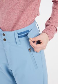 Lichtblauwe broek met een gladde textuur, voorzien van een ritszak, twee knopen aan de voorkant en een klittenbandsluiting om de tailleband aan te passen.