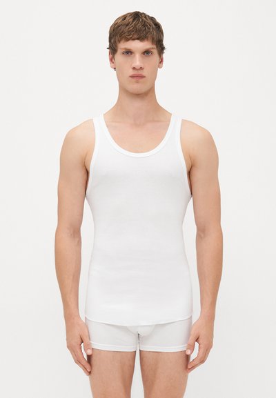 Junger Mann, der mit dem Gesicht nach vorne steht, trägt ein weißes geripptes Tanktop und weiße Boxershorts vor einem schlichten Hintergrund.