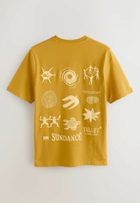 T-shirt jaune moutarde avec des symboles abstraits blancs, des figures dansantes, une tortue et le texte « 1999 Sundance Valley California Sounds. »