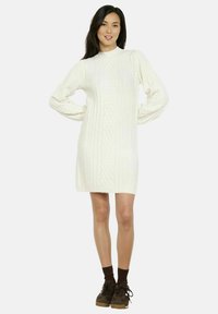 Vestito maglione bianco con collo alto, maniche lunghe e motivo a treccia testurizzata. Indossato con calzini marroni e sneakers.