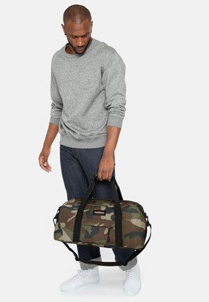 Bolsa de lona de camuflaje con correas negras, bolsillo con cremallera y parche de marca. Tela texturizada, forma redondeada, adecuada para viajes o para el gimnasio.
