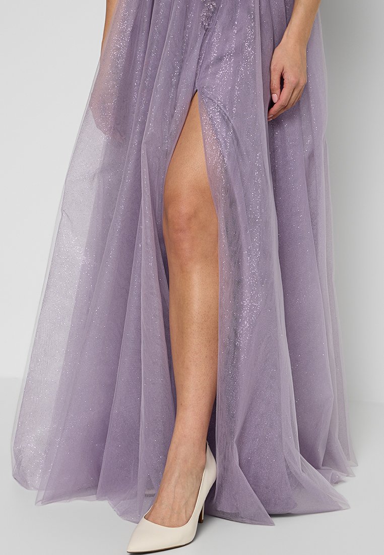 Suknia z lawendowego tiulu do ziemi z wysokim rozcięciem, zdobiona cekinami. Noszona w połączeniu z beżowymi szpilkami.