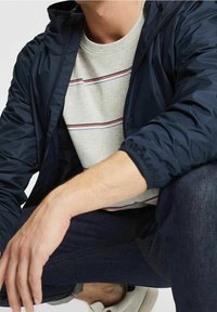 Veste navy au design ajusté, t-shirt gris rayé en dessous et jeans foncés. La veste possède des poignets froncés et une capuche.
