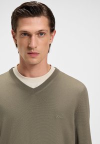 Junger Mann mit nach hinten gegeltem Haar, der einen beigen V-Ausschnitt-Pullover mit "BOSS"-Logo über einem hellen Creme-Hemd vor einem schlichten Hintergrund trägt.