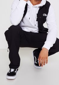 Mann in der Hocke, der schwarze Cargo-Hosen, schwarz-weiße Sneakers, einen weißen Hoodie und eine schwarze Weste mit dem The North Face-Logo trägt.