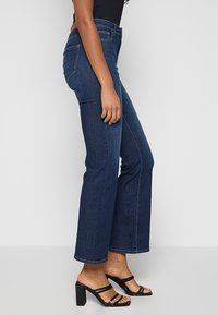 Tumed sinised flare-jeans, mis on valmistatud denimist, kõrge talje, viie tasku disain ja nähtavad õmblused õmblustes.