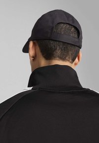 Sort zip-up jakke med høj krave, der har hvide accentstriber og en sort kasket. Teksturen ser glat og let struktureret ud.
