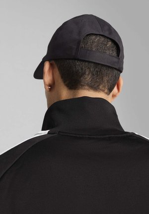 Sort zip-up jakke med høj krave, der har hvide accentstriber og en sort kasket. Teksturen ser glat og let struktureret ud.