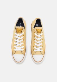 Żółte sneakersy z płótna z białymi sznurówkami, z gumowym noskiem i złotymi oczkami, z napisem "Converse All Star" na języku.