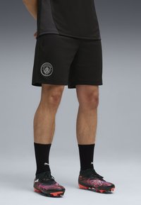 Une personne se tient debout en portant un short de sport noir avec le logo de Manchester City, des chaussettes noires et des crampons de football colorés, sur un fond gris.
