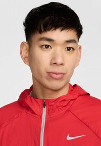 Veste légère rouge avec capuche, dotée d'une fermeture éclair intégrale et d'un logo Nike gris sur la poitrine. Texture lisse et design minimaliste.