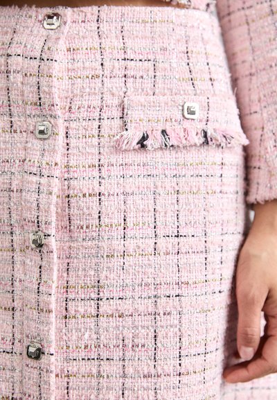 Nærbilde av rosa tweed-stoff med svarte, gull- og hvite tråder, sølvgeometriske knapper, en frynsete lomme og en hånd på siden.
