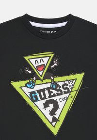 Schwarzes T-Shirt mit einem bunten Grafikdesign, das einen lächelnden dreieckigen Charakter auf übereinander liegenden grünen und weißen Dreiecken zeigt, sowie dem Text "GUESS".